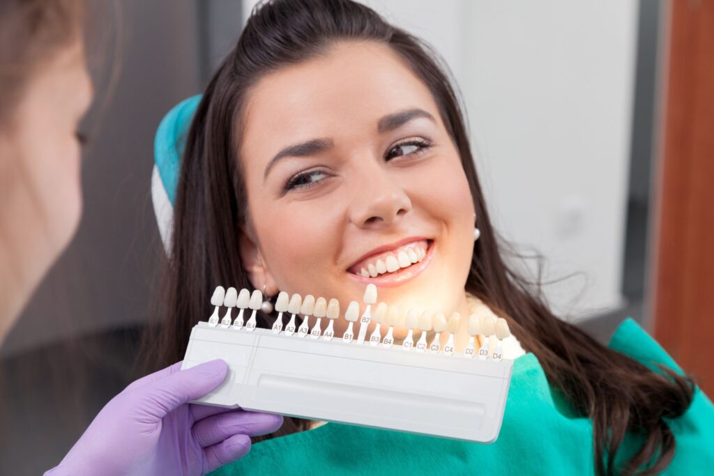 Woman with brown hair smiling at dentist holding shade guide to her teeth